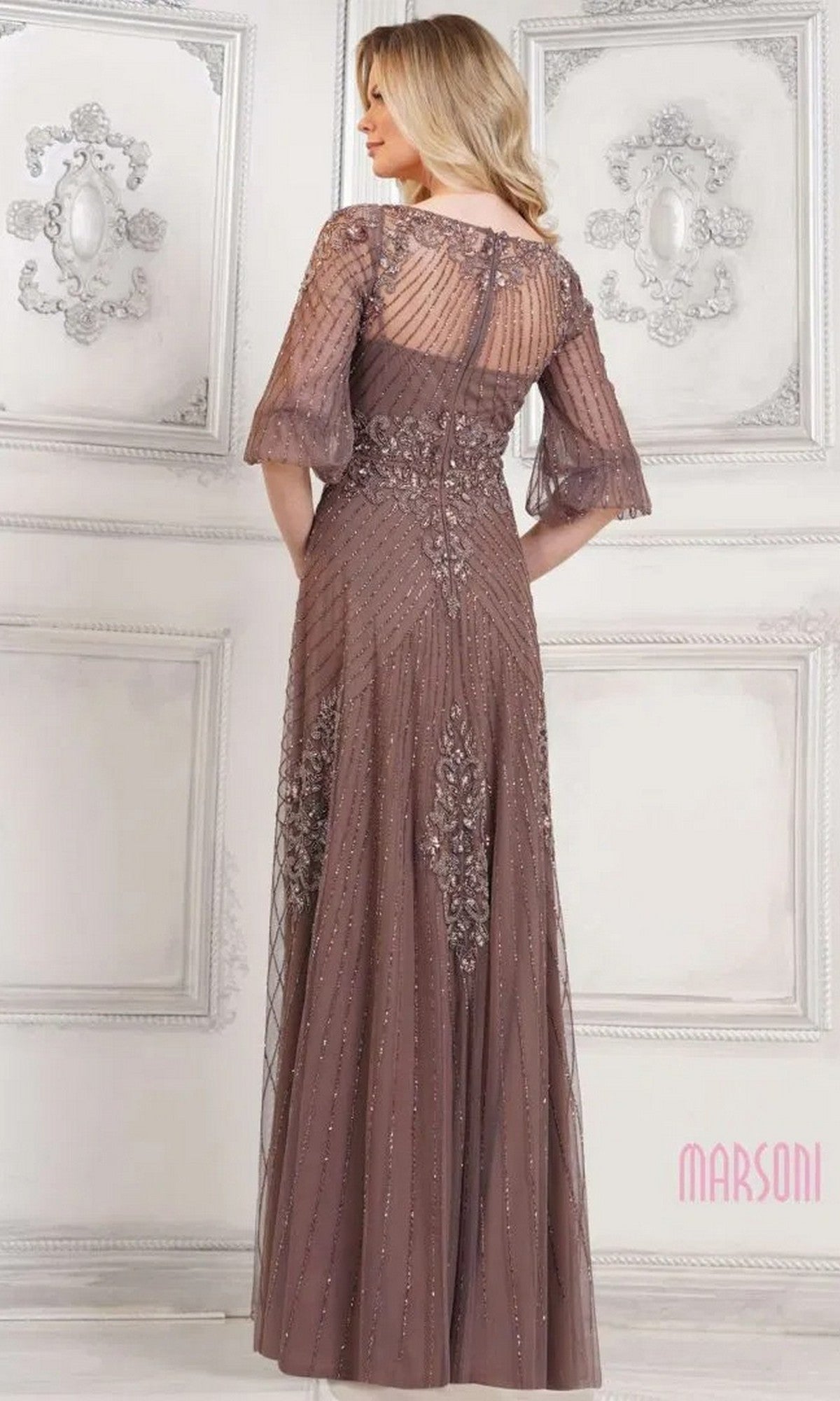 A woman stands with her back to the camera in an ornate room, wearing the Marsoni MV1323 long formal dress featuring sheer sleeves and intricate beaded embellishments.